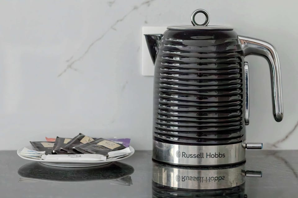 A close-up shot details a modern black and stainless steel electric kettle resting on a dark, glossy countertop. A small glass bowl filled with wrapped sweet treats sits nearby for the convenience of guests.