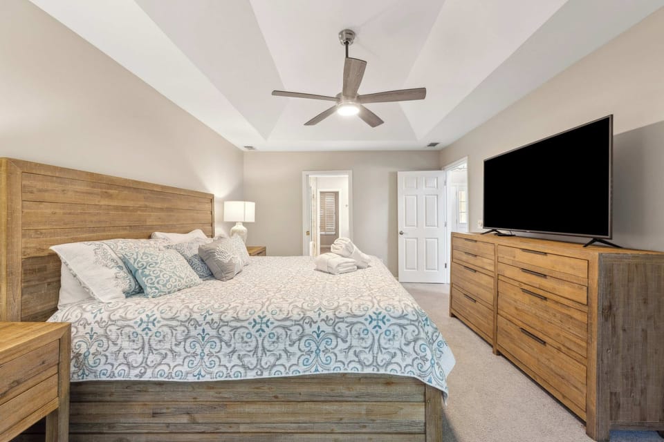 Rustic wood grain and cool patterned linens create a peaceful sanctuary under the high ceiling. A sleek fan and wide screen provide modern comfort, while gentle lamp glow highlights the organic textures of the matching furniture.