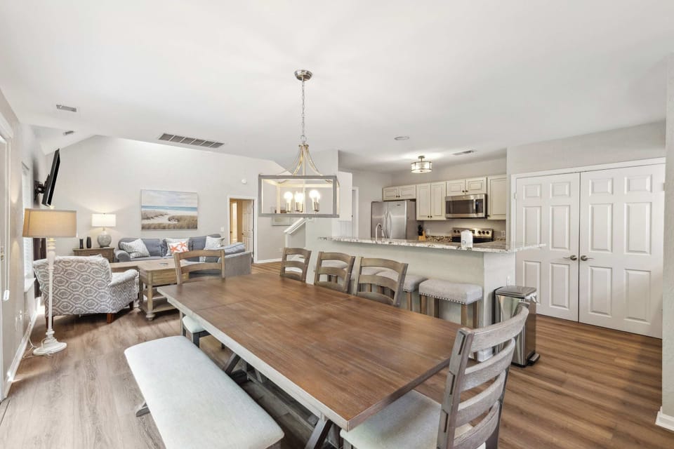 Golden wood tones and soft gray upholstery ground this open living area, where an elegant chandelier hangs above the dining table. Morning glow fills the space, connecting the cozy lounge to a modern kitchen perfect for shared meals.