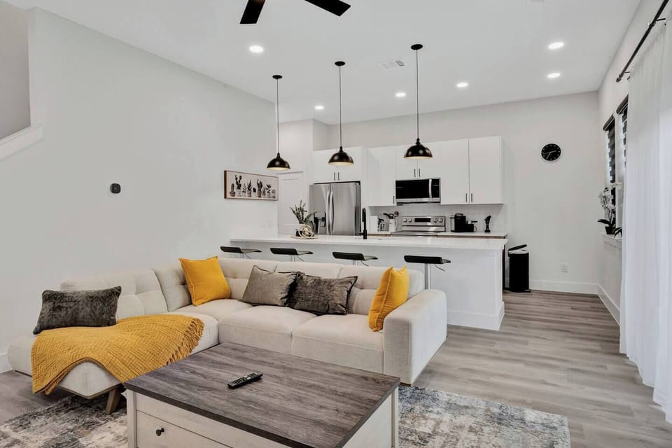 Bright and stylish living room with a cozy sectional sofa, smart TV, and open layout leading to the kitchen. Perfect for relaxing, watching movies, or enjoying time together.