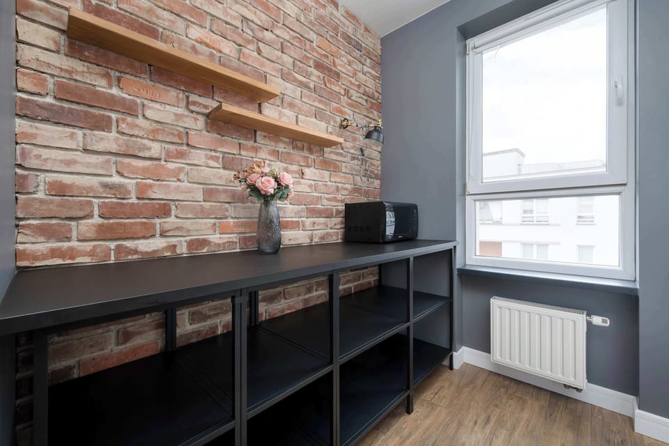 The brick accent wall is fitted with floating wooden shelves, creating a rustic storage display above the black shelving unit. A small vase of flowers sits on the lower counter near a white window frame.