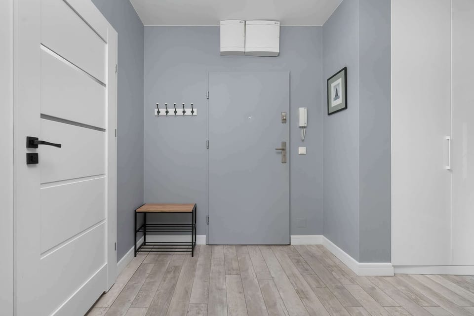 The entrance foyer features a light gray front door next to a simple wooden bench for seating. The gray walls and light flooring create a bright, neutral introduction to the apartment.
