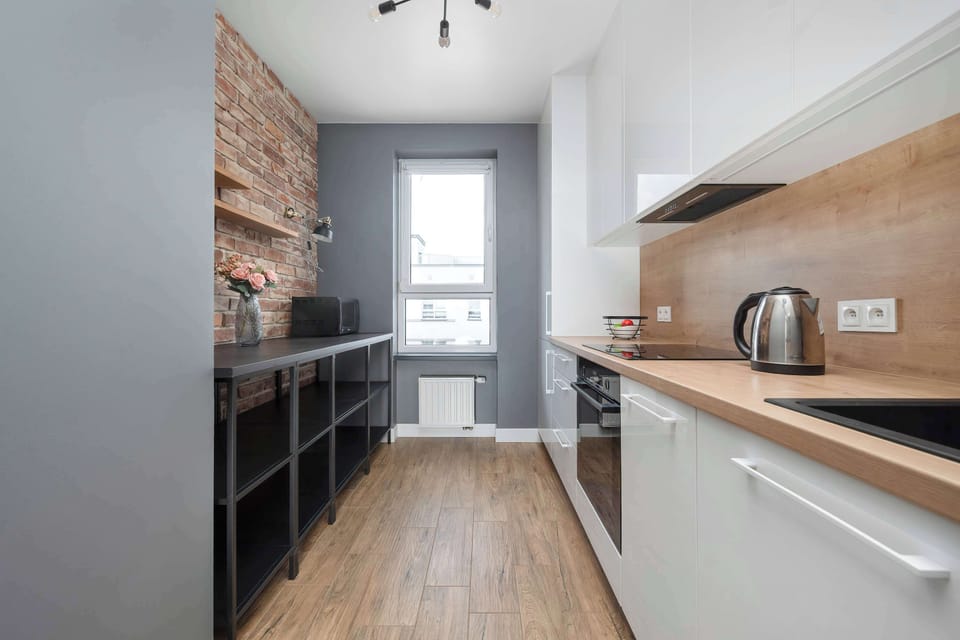 A galley-style kitchen walkway features white upper cabinets on one side and an open black shelving unit on the other. An exposed red-brick accent wall adds an industrial texture to the long space.
