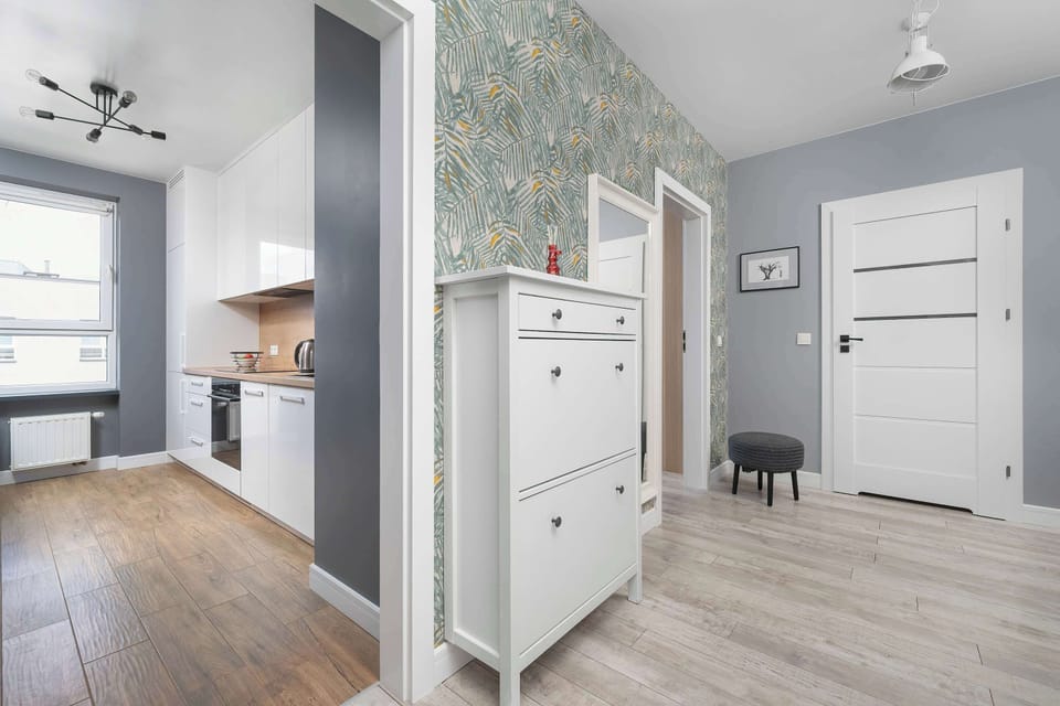 The apartment entrance looks from the kitchen into a hallway decorated with patterned floral wallpaper. A white shoe storage cabinet sits on the light wood-plank floor next to a white door.