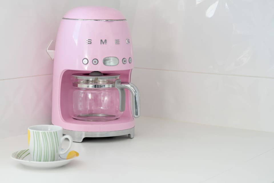 A retro-style pink coffee machine placed on a spotless white countertop. Small accessories beside it add a decorative touch.