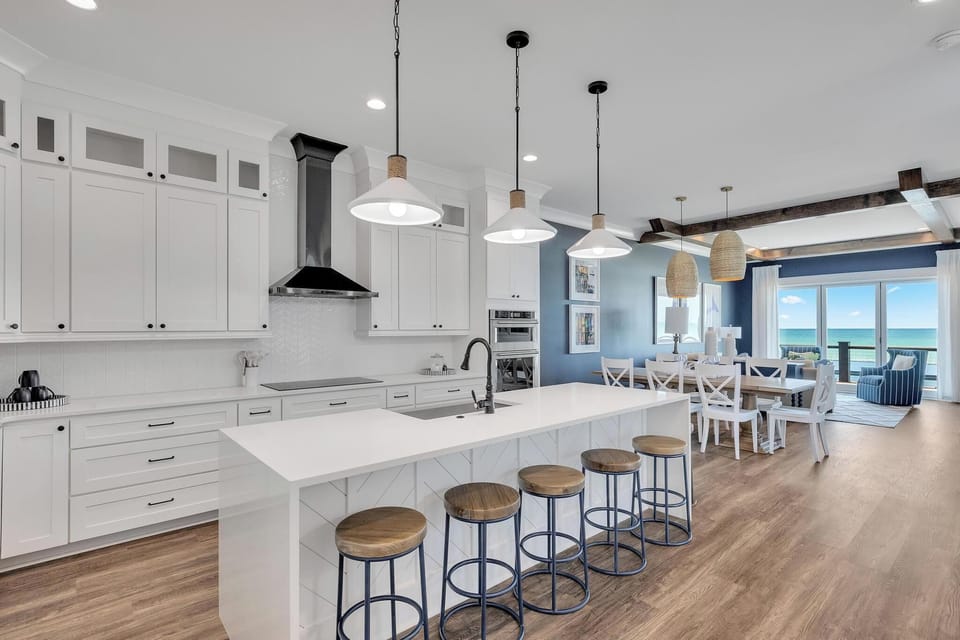 Step into your fully equipped kitchen with gleaming white cabinetry, expansive island seating, and an open layout flowing to the dining area where stunning ocean views await through floor-to-ceiling windows.