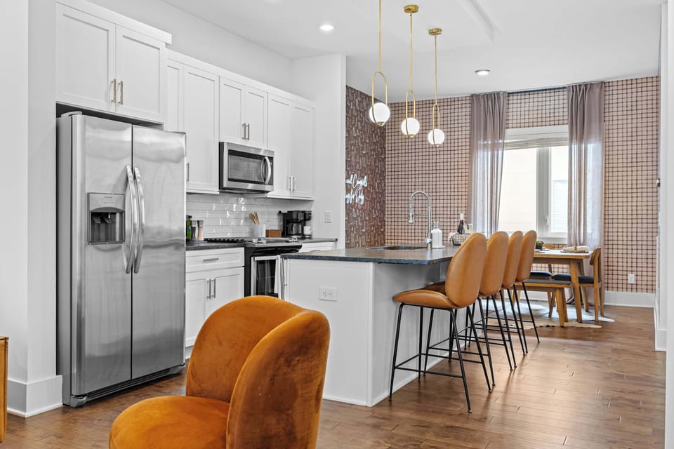 White shaker cabinets, stainless appliances, subway tile backsplash, and a dark granite island with seating for four flow into a dining nook wrapped in western-print wallpaper and gold pendant lights. -