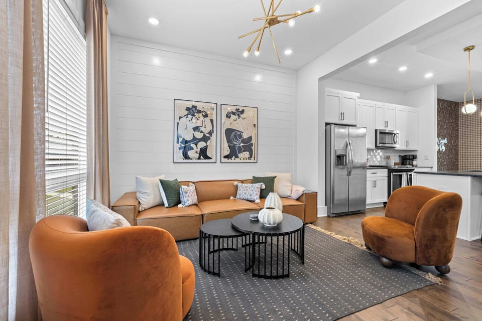 The open layout connects the living room to the kitchen seamlessly. Leather sofa, velvet accent chairs, and stainless steel appliances all visible from one inviting space.