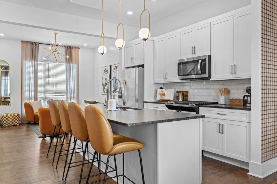 A large kitchen island with seating for four sits beneath gold globe pendant lights, with white shaker cabinets, subway tile backsplash, and stainless appliances just steps from the living room.