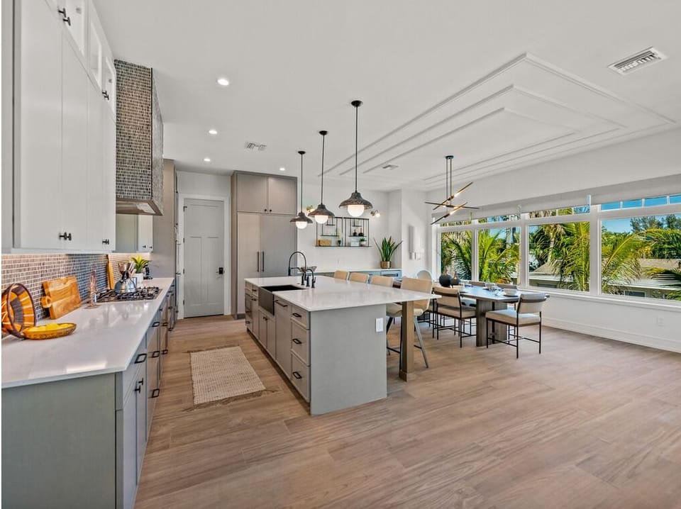 Gather around this bright open kitchen with sleek countertops, modern appliances, and a large island perfect for cooking together. Sunlight streams through expansive windows framing tropical palm views, while the adjoining dining space invites memorable me