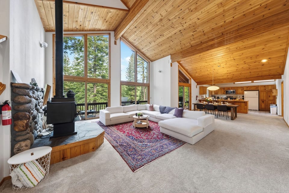 Sun-drenched, open-concept living room featuring floor-to-ceiling windows, soaring vaulted wood ceilings, a cozy wood-burning stove, and access to the outdoor deck.