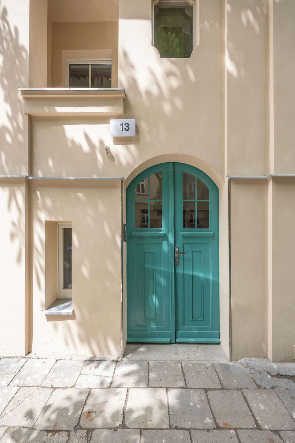 A building exterior detail focuses on a classic arched entrance door painted in a vibrant teal color. The stone pavement and beige facade reflect traditional European townhouse architecture.