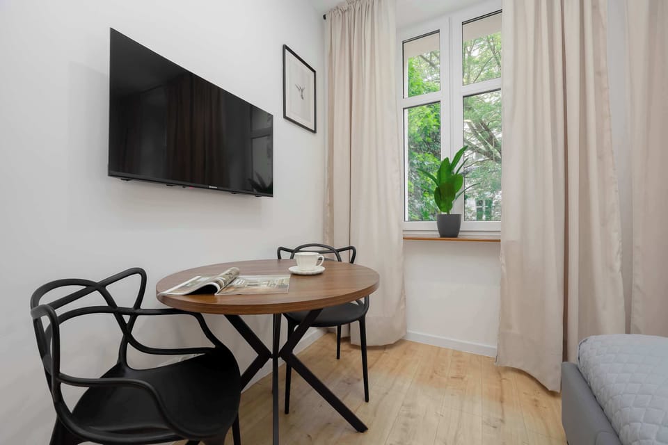 The cozy dining area features a wooden table paired with two modern black designer chairs. A window brings in natural light, and a small framed print on the wall completes the simple decor.