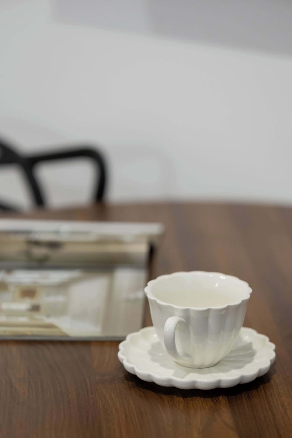 A tabletop macro shot focuses on a delicate white ceramic teacup and saucer resting on the dark wood surface. A magazine lies blurred in the background, creating a peaceful morning vibe.