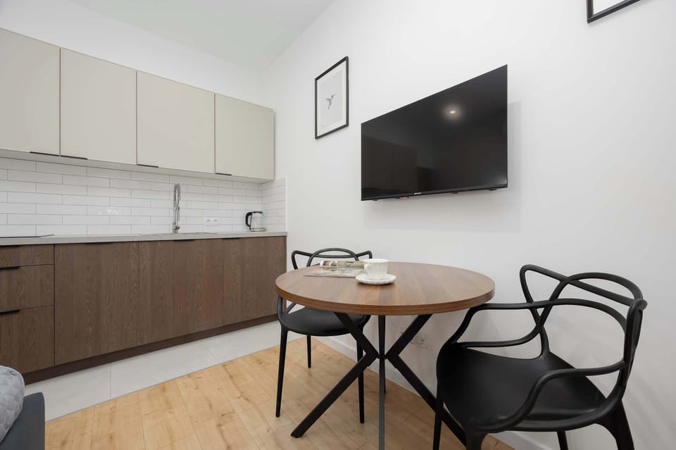 The dining setup captures the round wooden table and two black mesh chairs in front of the kitchen wall. The open-plan design maximizes the utility of the studio apartment.