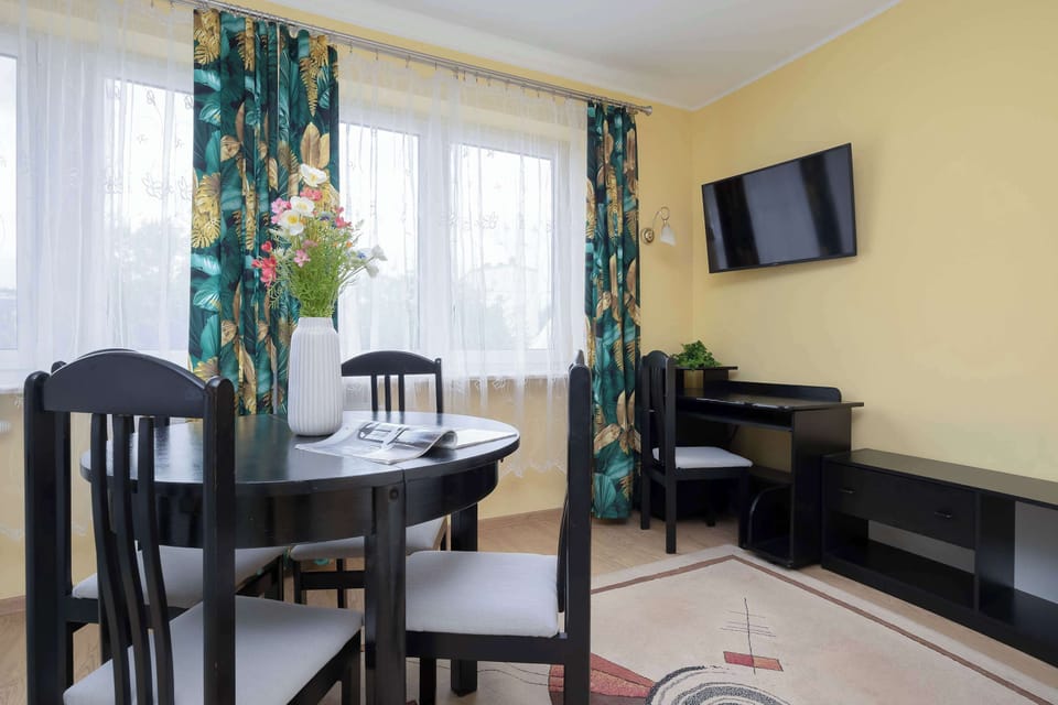 A bright and cozy living room with a dining area, large windows dressed with colorful curtains, a wall-mounted TV, and a compact workspace.
