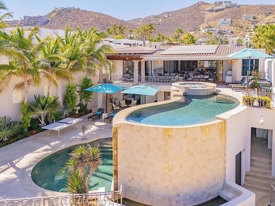 A luxurious desert retreat nestled in the hillside, featuring tiered pools, lush tropical gardens, and expansive terraces with dramatic mountain and residential views.