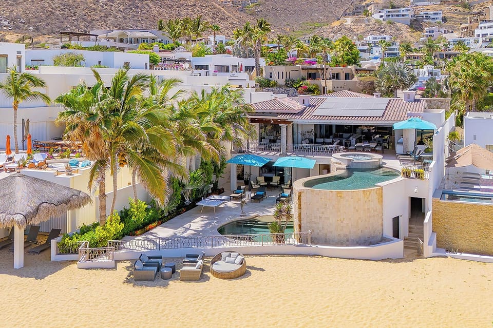 Hillside beachfront villa showcasing multi-level pools, private sandy beach access, tropical palms, and sunny outdoor lounging areas in an exclusive coastal neighborhood.