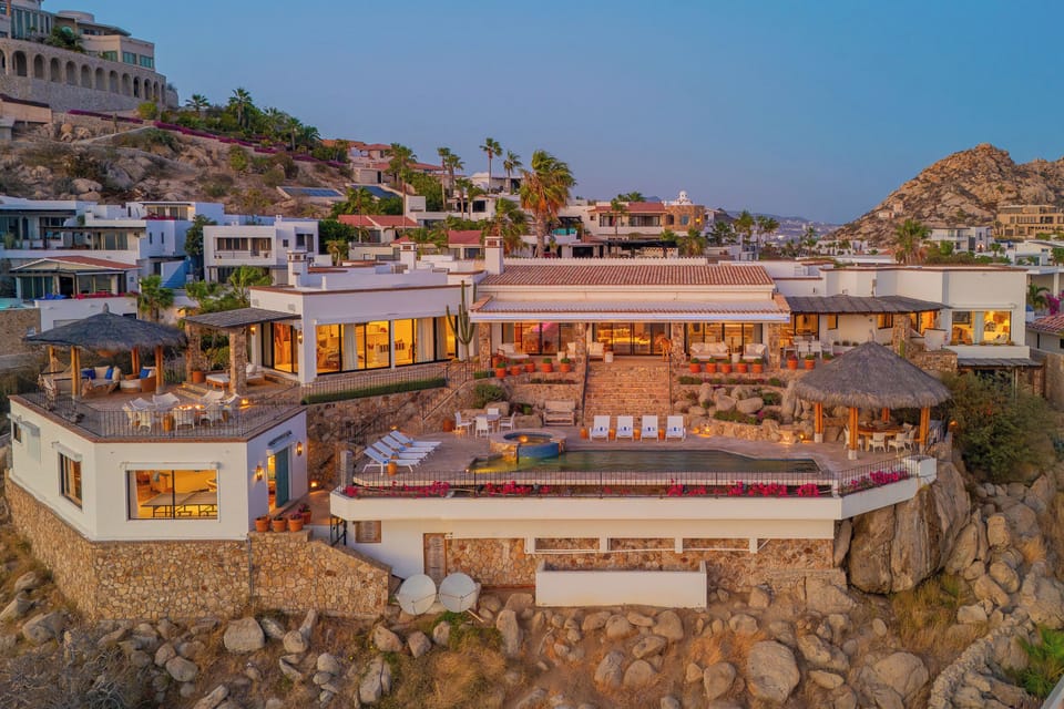 Luxury hillside villa perched on dramatic rocky coastline, featuring multiple levels with pools, terraces and thatched-roof palapas overlooking the sea.