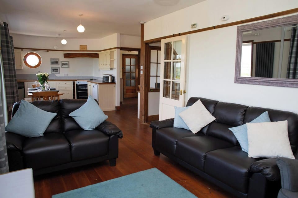 Living area with black leather sofas, pale cushions, wooden floor and kitchen beyond