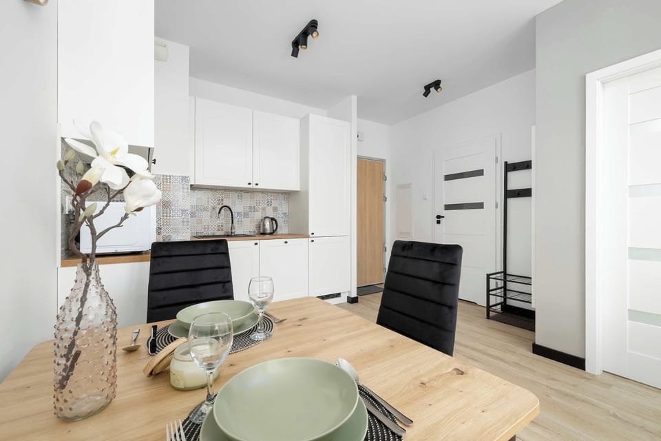 A wooden dining table is beautifully set with plates and elegant glassware, positioned next to a compact kitchen zone. Black chairs offer a bold contrast to the bright, white cabinets in the background.