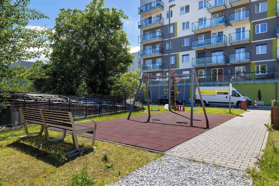 An outdoor children's playground features a swing set and a bench surrounded by a secure fence and green trees. A modern multi-story residential building stands prominently in the background.