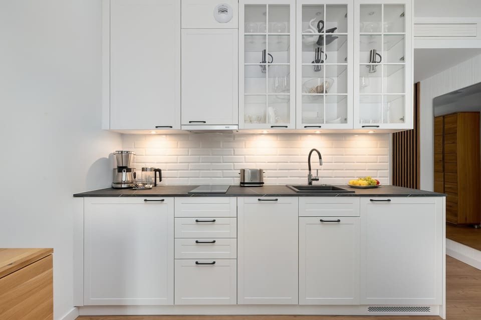 A minimalist kitchenette is equipped with elegant white upper and lower cabinets, some of which feature glass fronts. A sink and an electric kettle are neatly placed on the dark countertop.