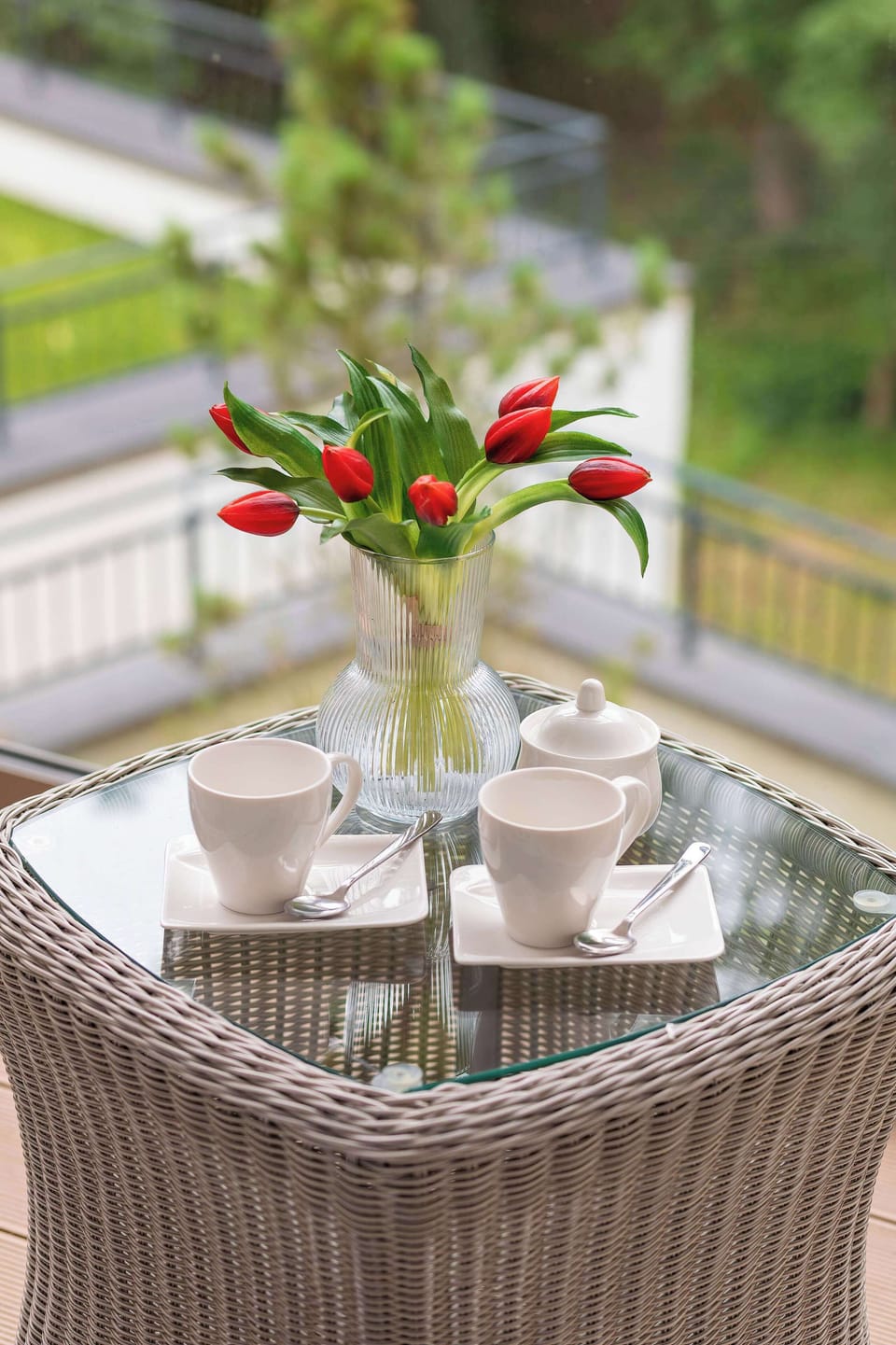 A close-up of a glass balcony table holding white cups and a vase with red tulips. A woven armchair invites relaxation in the fresh air.