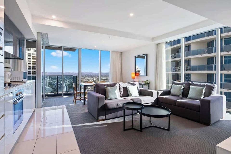 Relax in this light-filled, open-plan living area featuring comfortable grey sofas, sleek black nesting coffee tables, an adjacent modern kitchen, and expansive floor-to-ceiling windows leading to a private balcony with elevated city views.