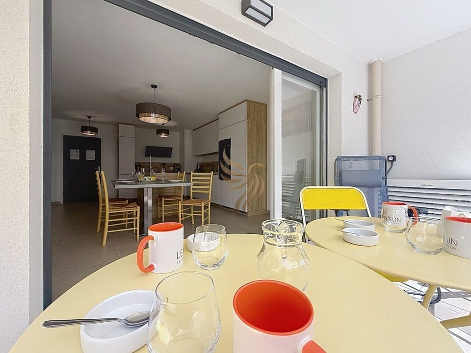 Petit-déjeuner au soleil : mugs orange sur la table jaune, la baie vitrée s`ouvre sur une cuisine claire et une grande table prête à accueillir un repas chaleureux entre amis.