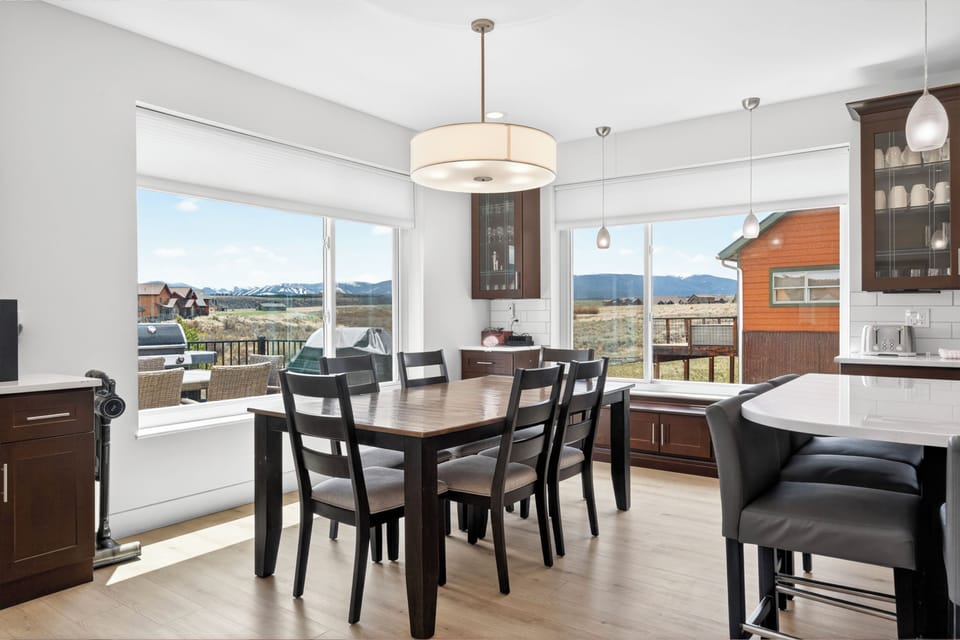 Gather around the spacious dining table where panoramic mountain views frame every meal. Sunlight pours through floor-to-ceiling windows, highlighting the open layout connecting your dining space to the sleek, modern kitchen and comfortable living area.