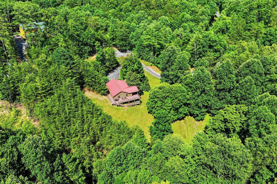 An aerial view captures a charming cabin with a red roof nestled in dense forest, offering complete privacy surrounded by lush green woodland and winding access roads.