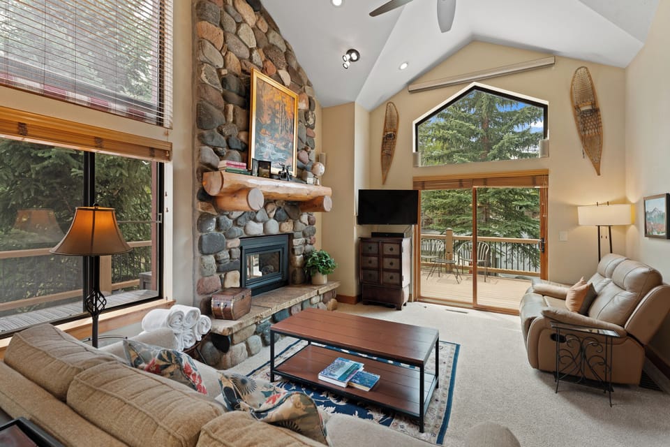 Relax in your mountain-retreat living room featuring soaring vaulted ceilings, a stunning stone fireplace, and private balcony access with forest views to complete your alpine escape.