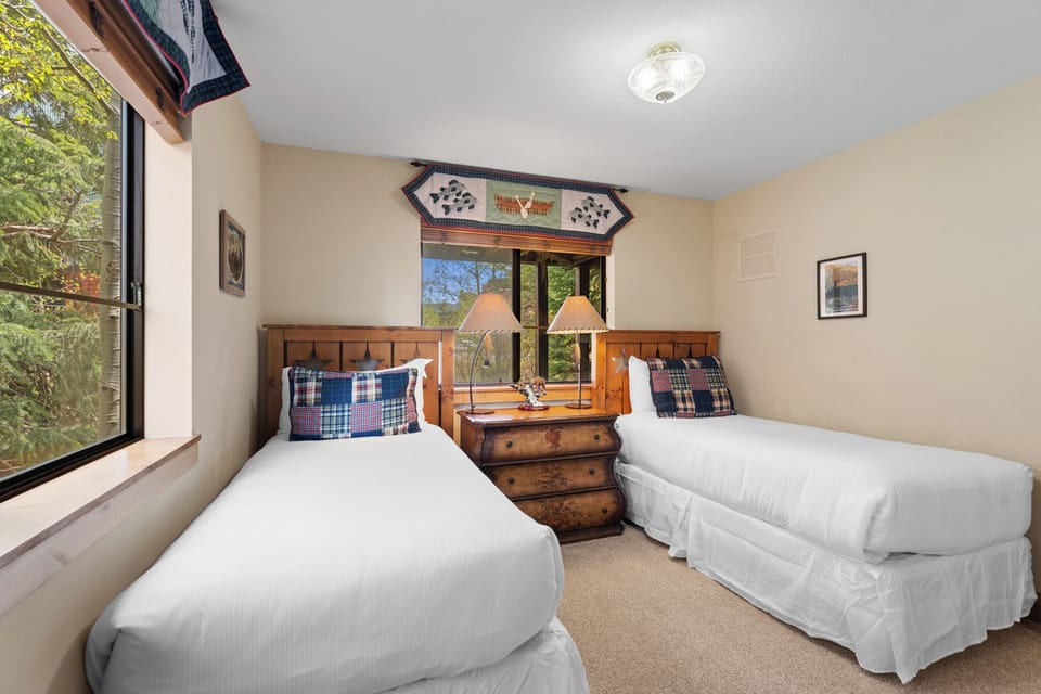 Unwind in this cozy bedroom with twin beds dressed in crisp linens and charming plaid accents. Natural light streams through large windows revealing peaceful garden views.
