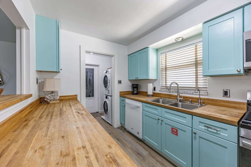 Kitchen with light blue cabinets, wooden countertops, and a stacked washer and dryer in the adjoining laundry room.