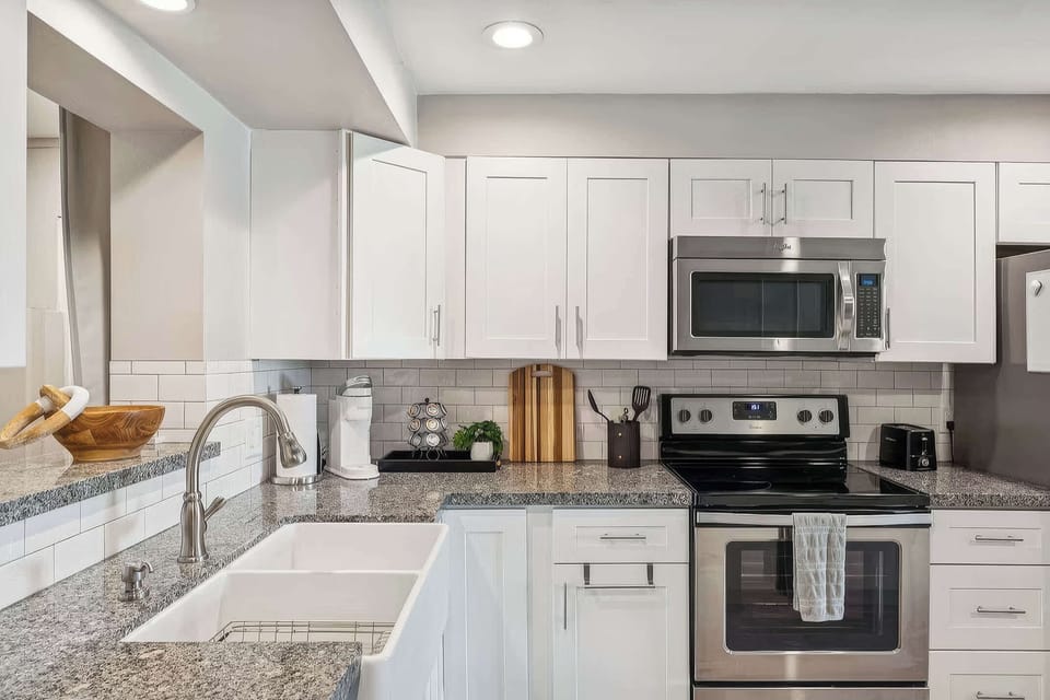 The kitchen impresses with a large granite island with seating for 2, farmhouse sink, white shaker cabinets, tile backsplash, Keurig coffee maker, and full stainless steel appliance suite.