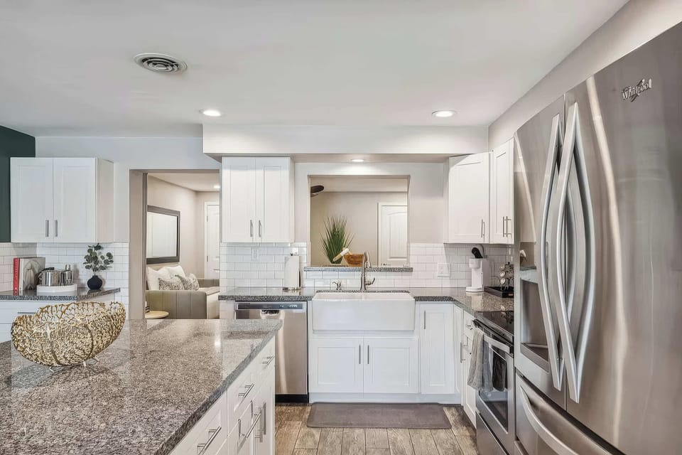 The kitchen impresses with a large granite island with seating for 2, farmhouse sink, white shaker cabinets, tile backsplash, Keurig coffee maker, and full stainless steel appliance suite.