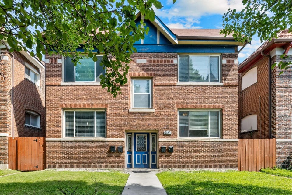 The daytime exterior shows the brick multi-unit building, front lawn, central walkway, and blue entry door.