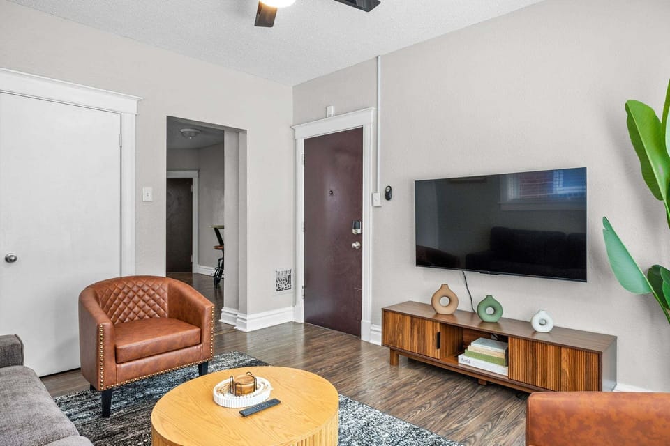 This reverse living-room view shows the wall-mounted TV, media console, seating, front door, and hall opening.