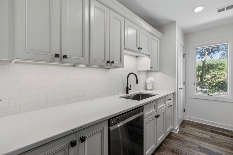 Your sleek, modern kitchen features pristine white countertops, stylish black fixtures, and a bright window overlooking lush greenery—perfect for preparing your favorite meals in comfort and style.