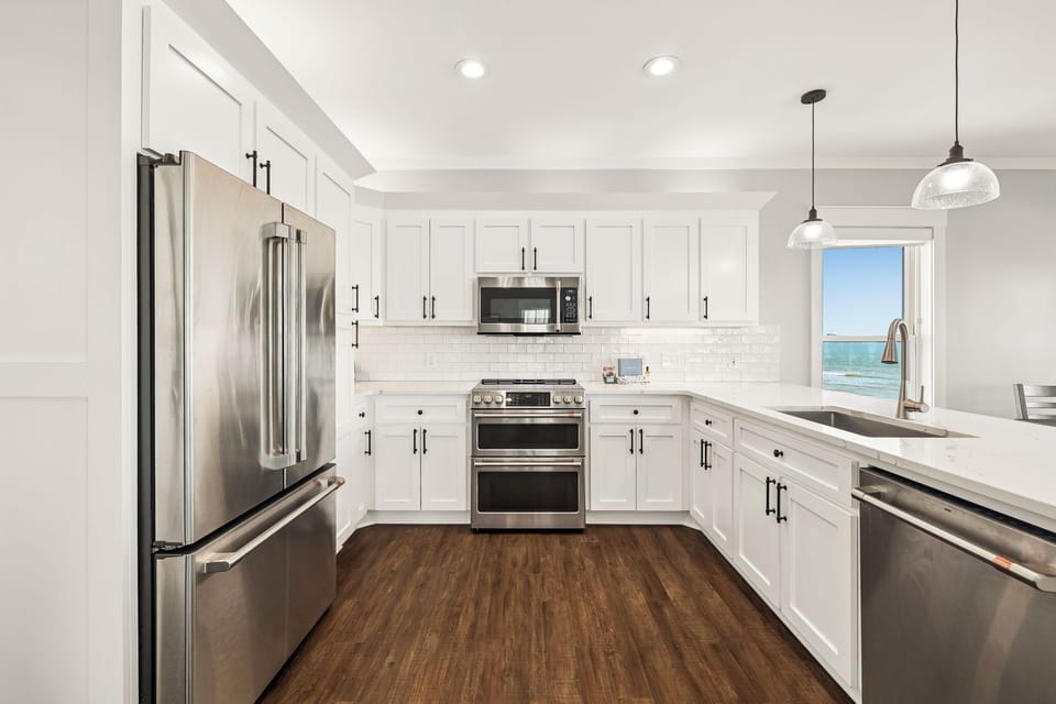 Step into your bright, fully equipped kitchen with crisp white cabinetry, stainless steel appliances, and stunning ocean views framed by the window—perfect for preparing meals while soaking in the coastal scenery.