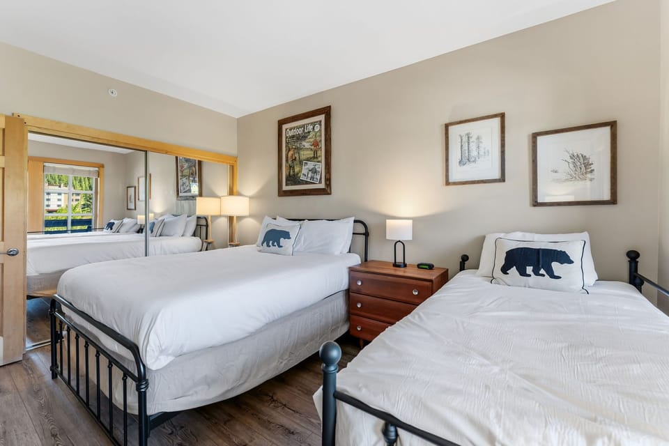 Sink into crisp linens with charming bear-themed pillows in this inviting bedroom. Two beds offer flexibility, while warm wood floors and nature-inspired artwork create a cozy mountain retreat.