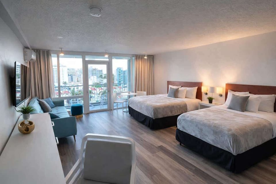 Spacious room featuring two comfortable queen beds with plush linens and modern finishes. Floor-to-ceiling windows fill the space with natural light and lead to a private balcony — perfect for relaxing after a day out exploring.