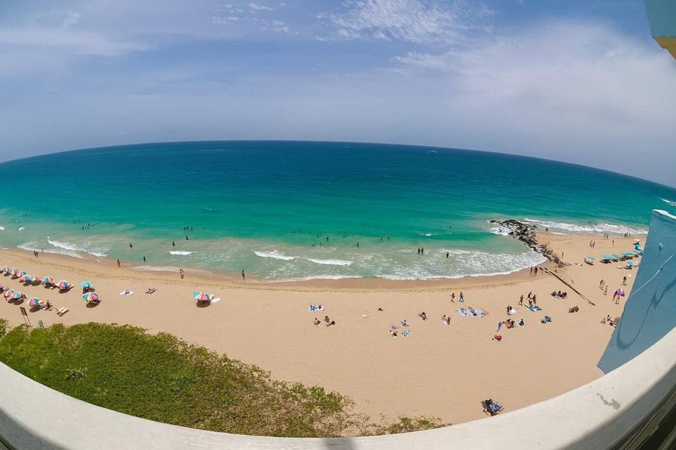 Panoramic beach view with turquoise waters, soft golden sand, and colorful umbrellas — a perfect oceanfront escape right outside your window.
