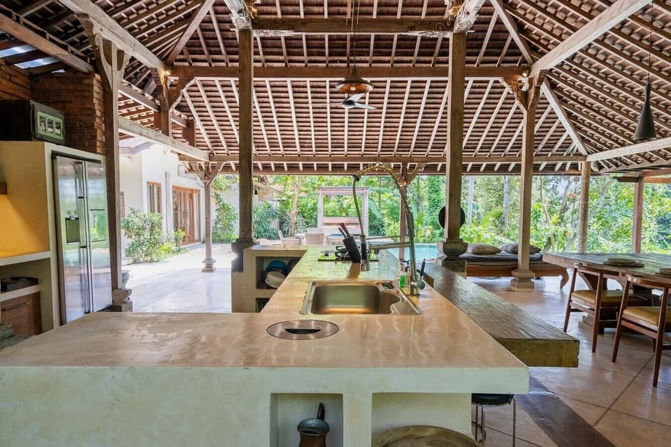 A spacious dining pavilion with a large wooden table, set under a traditional thatched roof. The area is open to the surrounding gardens, offering a perfect spot for outdoor dining.
