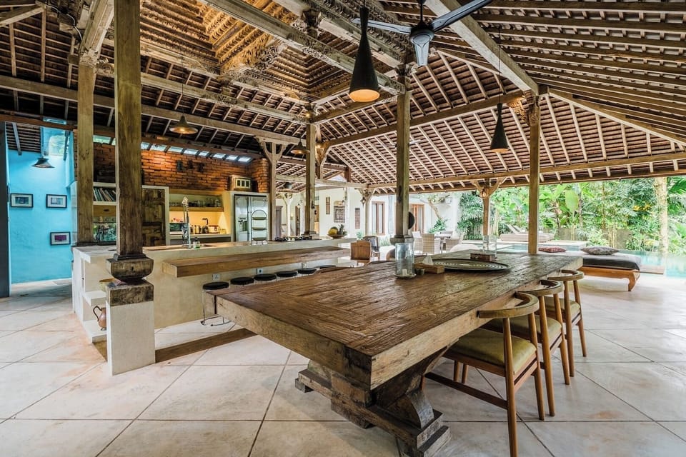 A rustic dining area with a large wooden table and benches, located under a thatched roof. The space is ideal for enjoying meals with family and friends in a tropical setting.