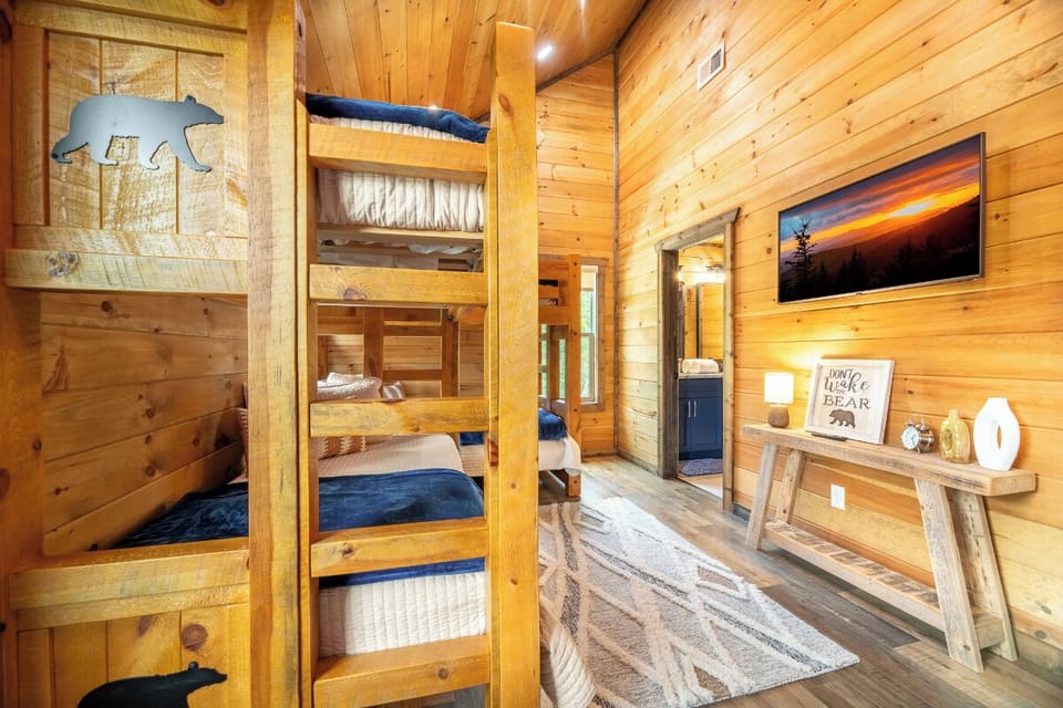 Your cozy cabin bedroom features rustic bunk beds with bear-carved details, warm wood tones, and charming woodland accents that invite restful nights under the vaulted pine ceiling.