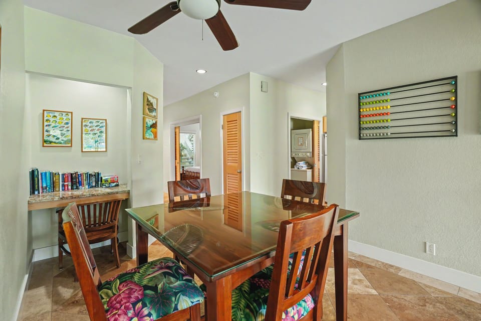 Your dining space features a glass-top table surrounded by tropical-print chairs, with a ceiling fan above and a cozy reading nook lined with books to enjoy during your stay.