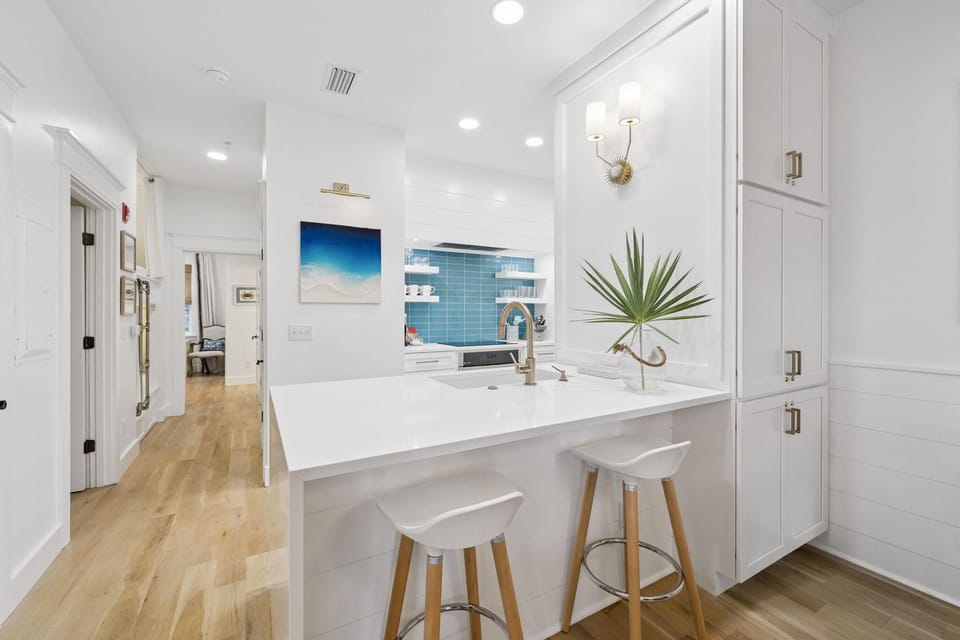 Two white stools with wooden legs sit beneath a quartz countertop accented by a glass vase and brass faucet.