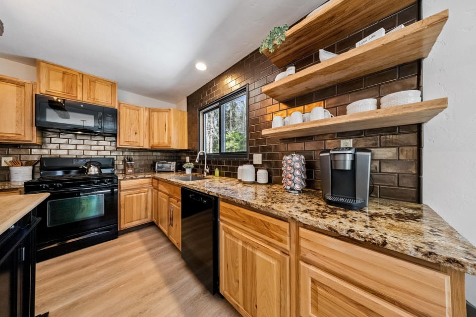 Granite countertop stretches beneath two natural wood floating shelves filled with white dishware and greenery.
